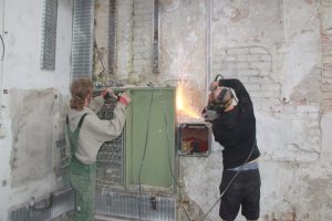 zwei Männer arbeiten vor einer unverputzen Wand - einer sägt - es sprühen Funken, einer arbeitet an einem alten Sicherungskasten